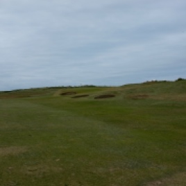 The view from the 16th fairway. Note the three bunkers, starting with two on the right-hand side of the fairway and a third set into the bank to the front left of the green. The staggered arrangement of these bunkers, set against the left to right dog-leg shape of the hole gives an advantage to those who can safely drive down the right-hand side of the fairway and avoid the first two of these hazards. The further left you go with your tee shot the more the bunker guarding the left half of the green comes into play.